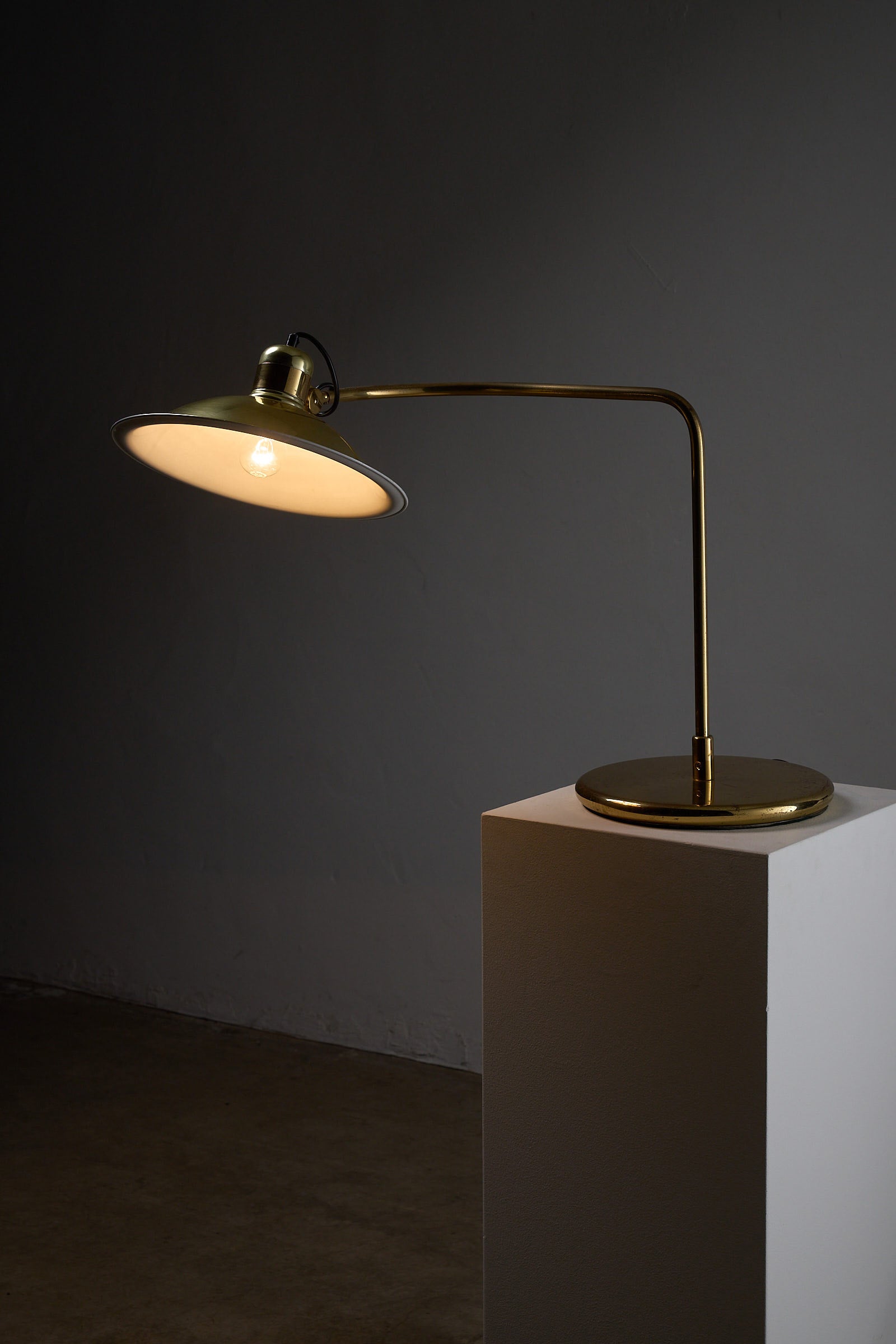 Cosack Brass Table Lamp with Curved Arm on a pedestal in a dark setting, showcasing its elegant design.