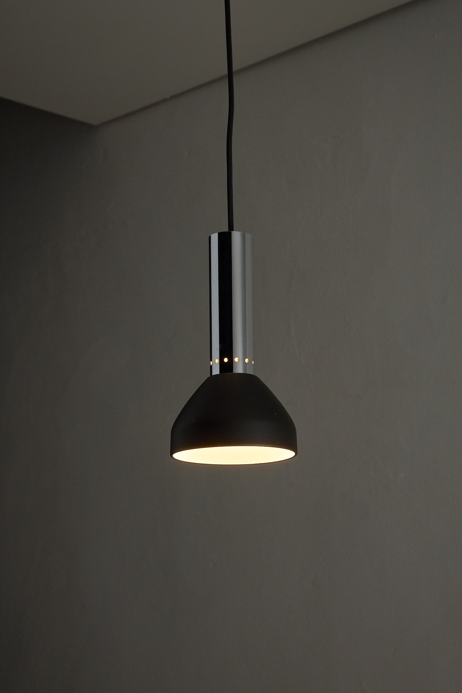 Chrome and Black Pendant Lamp with Perforated Detail hanging against a grey wall, providing warm focused downlight.