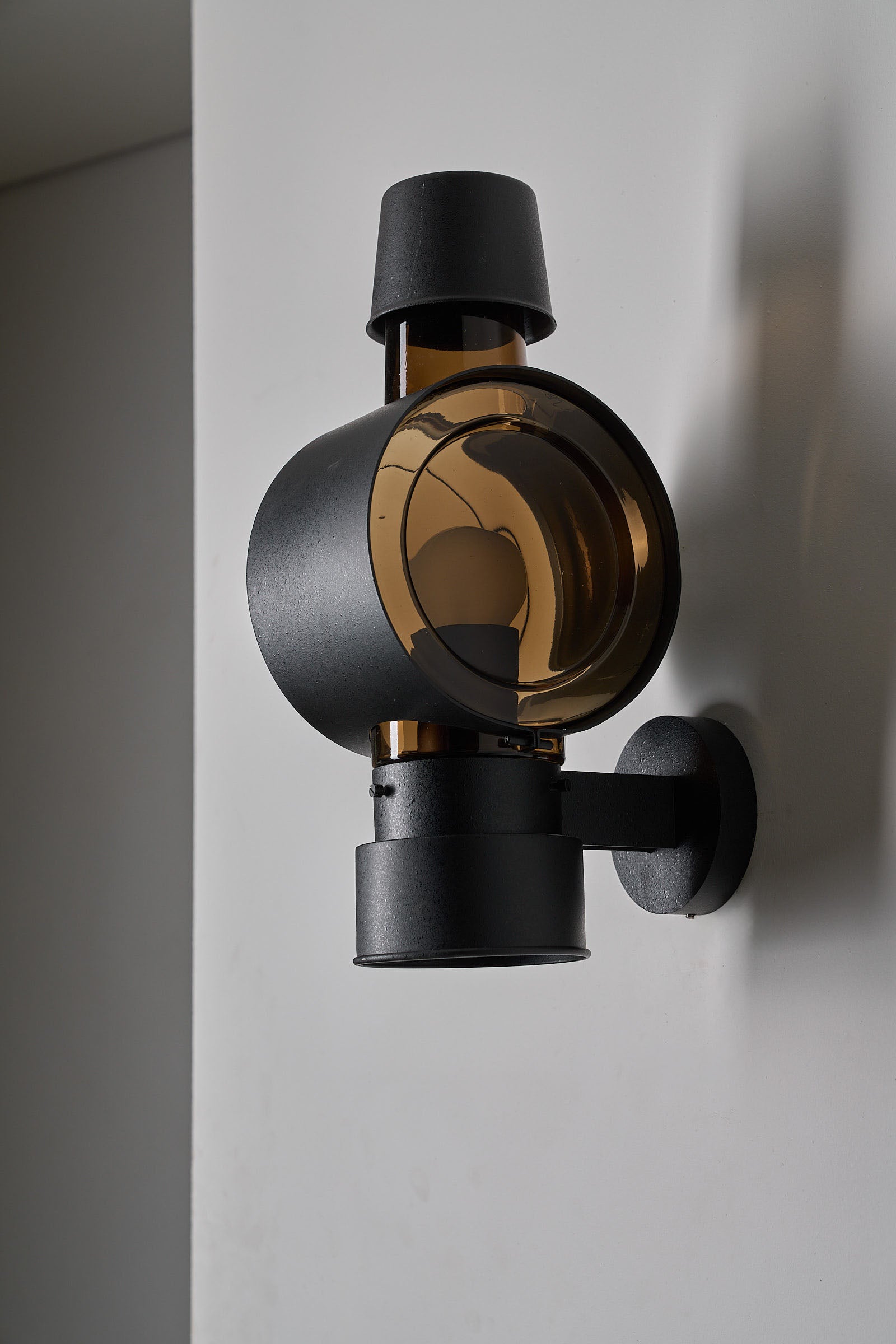 Bega Boom Outdoor Sconce Cylindrical Amber Glass on a wall, featuring amber-brown glass and textured dark metal.