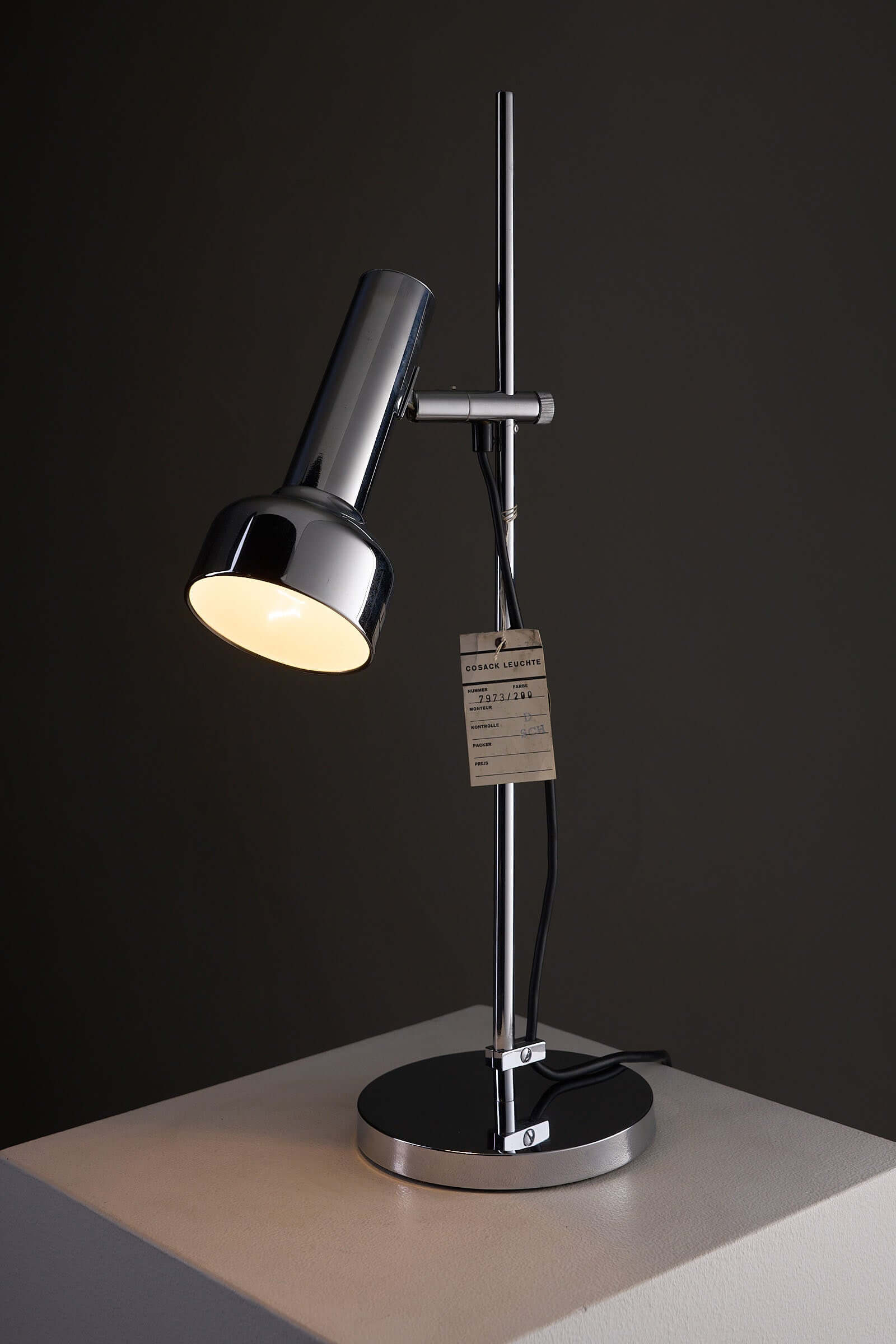 Adjustable Chrome Table Lamp Model 7973 by Cosack on a pedestal with warm light and manufacturer tag hanging.