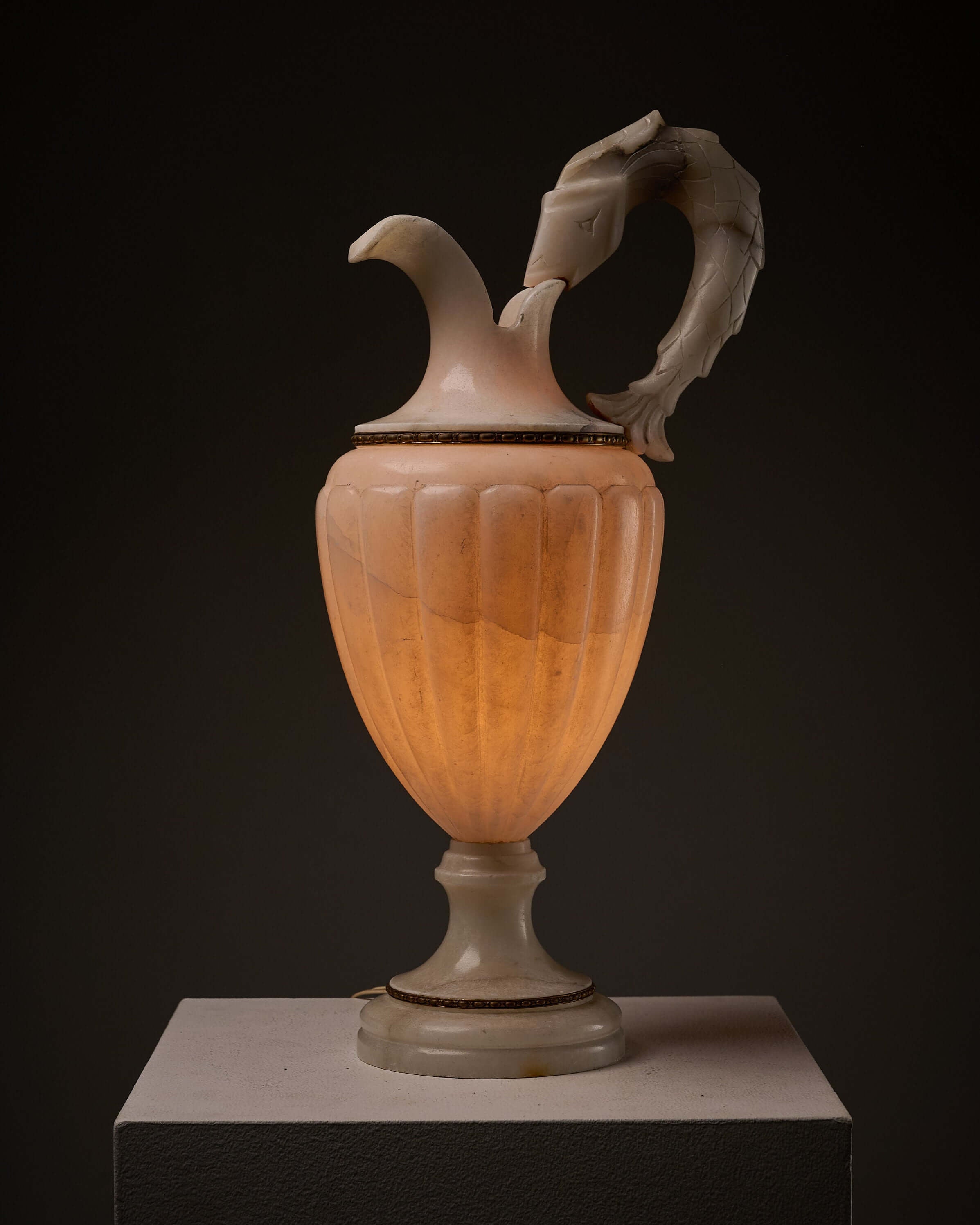 Carved Alabaster Urn Lamp with Stylized Fish Handle displayed on a pedestal against a dark background.