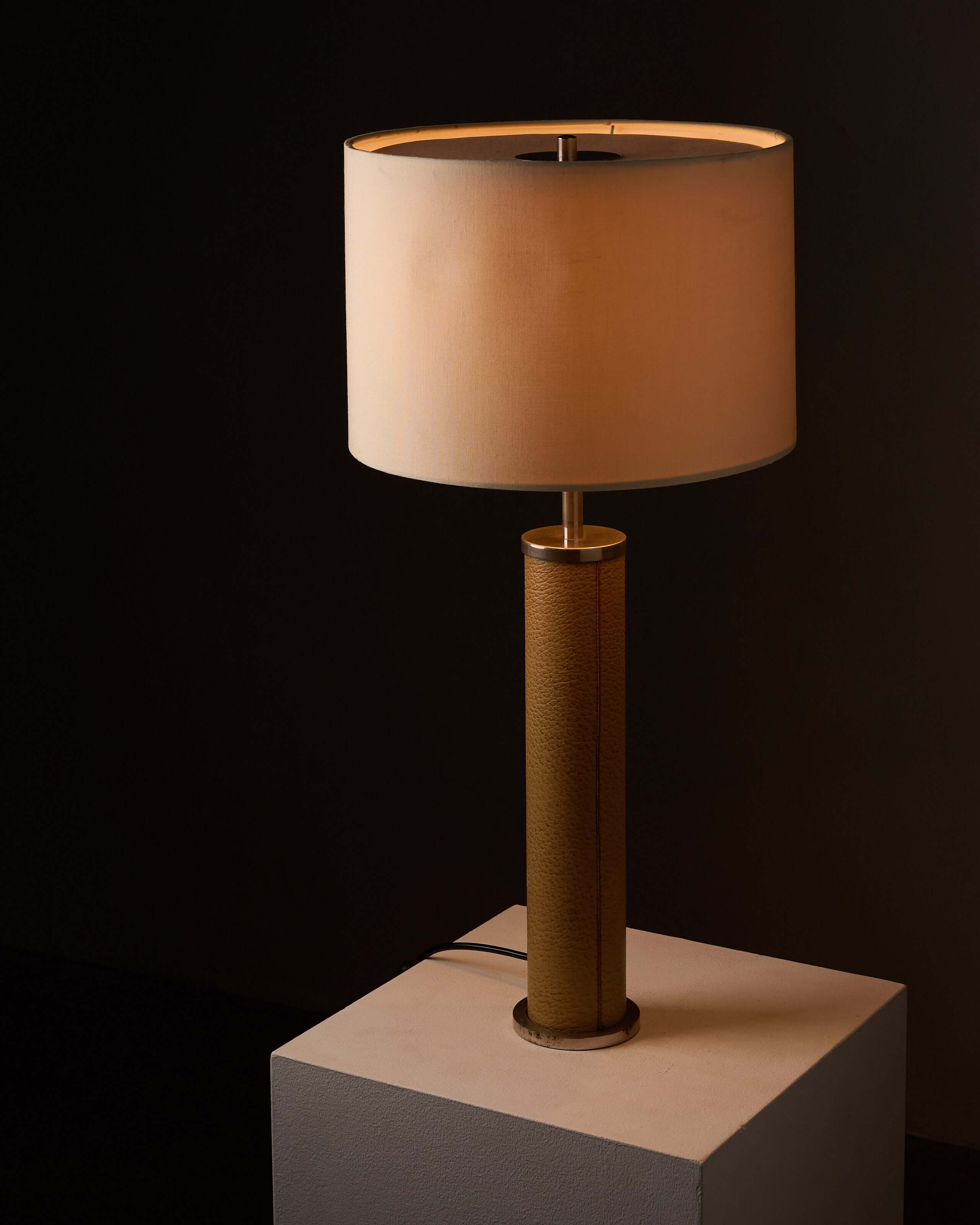 Cylinder Table Lamp in Stitched Natural Leather with tan leather base and neutral fabric shade providing ambient light.