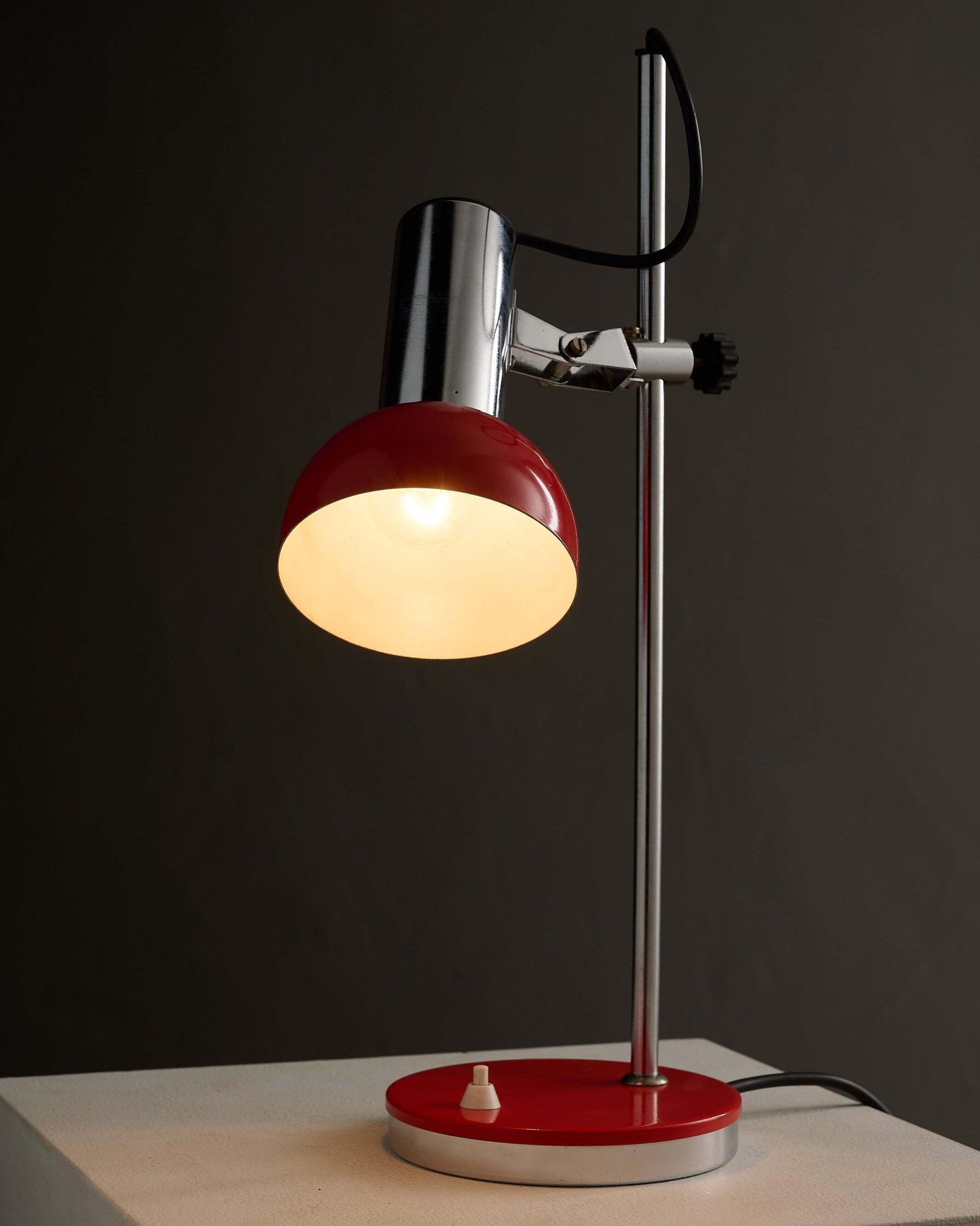 Adjustable chrome red table lamp with red shade and weighted base on a pedestal.