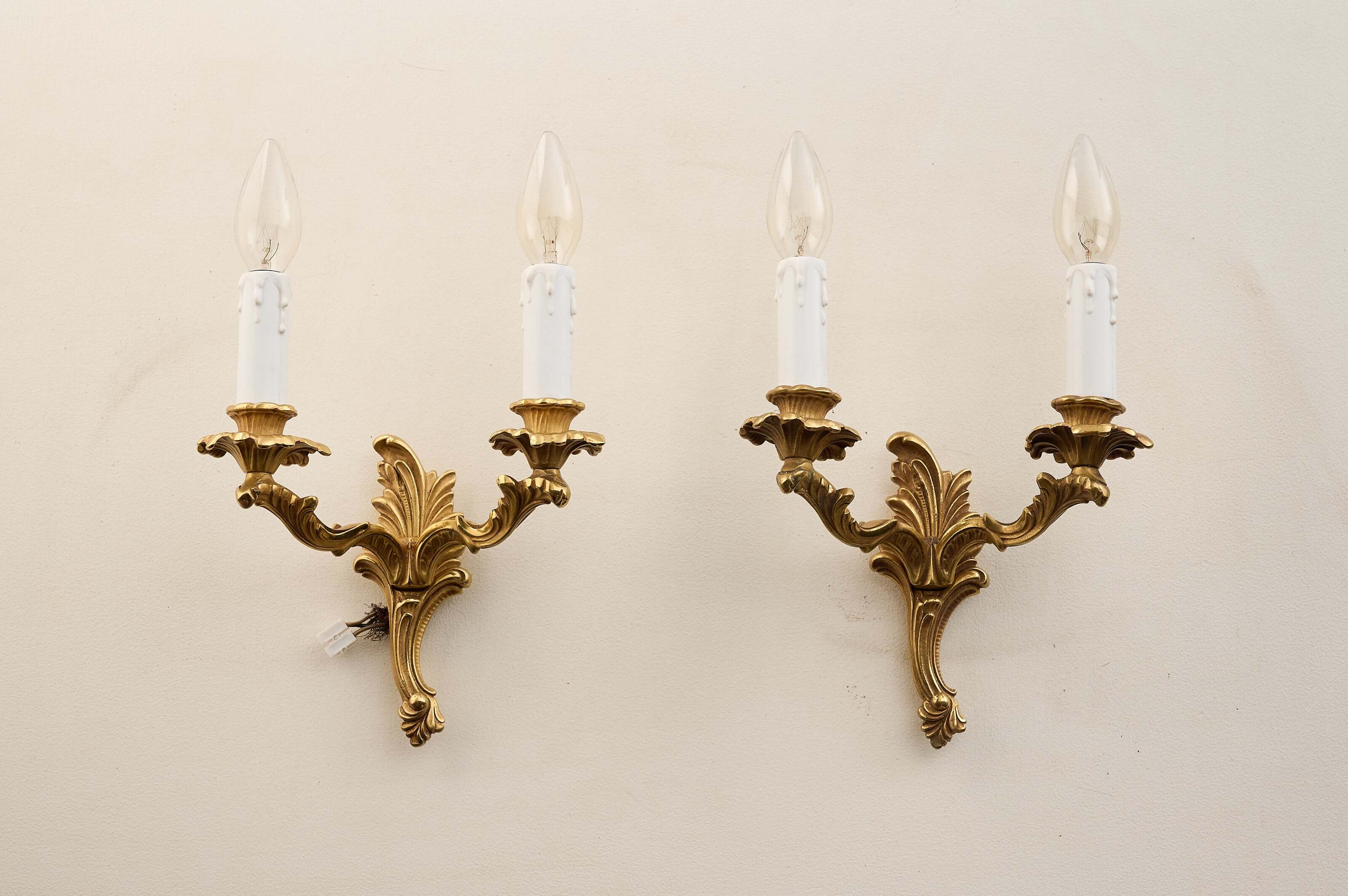 Pair of Louis XV style brass wall sconces featuring decorative arms and faux-candle sleeves.