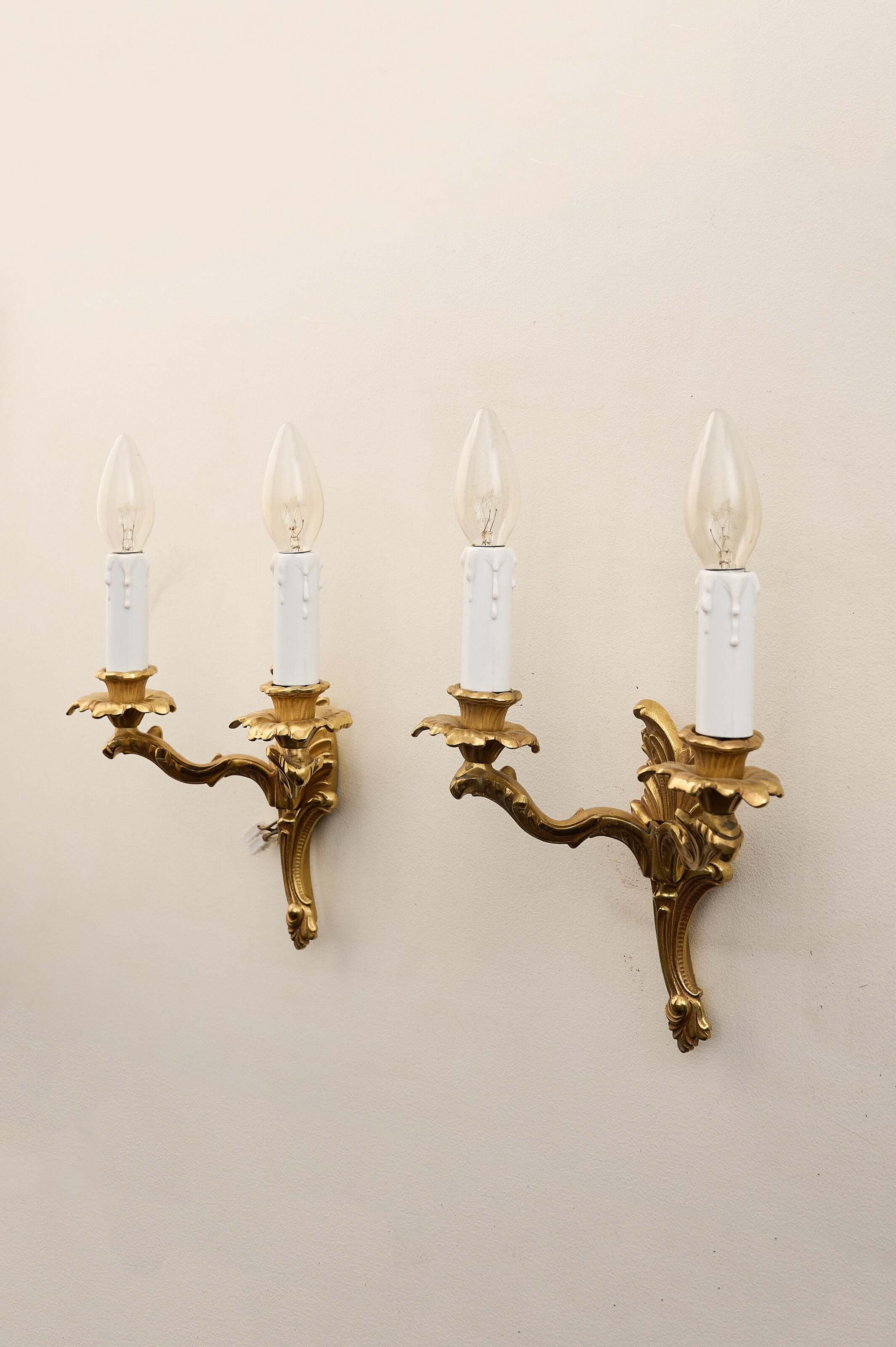 Pair of Louis XV style brass wall sconces with double arms, floral bobèches, and white faux-candle sleeves.