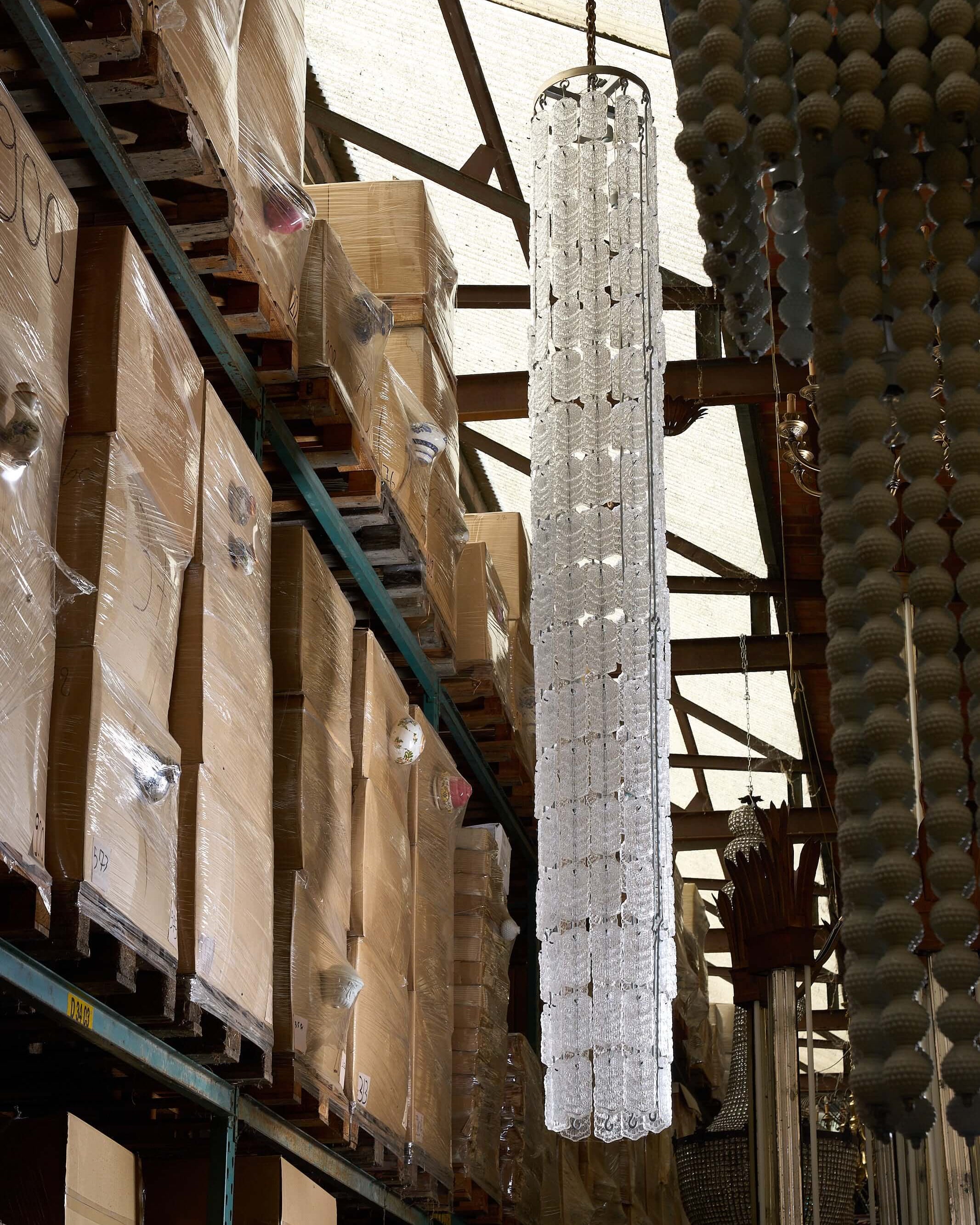 XXL Glass Column Chandelier displayed in a warehouse among stacked boxes and other lighting fixtures.
