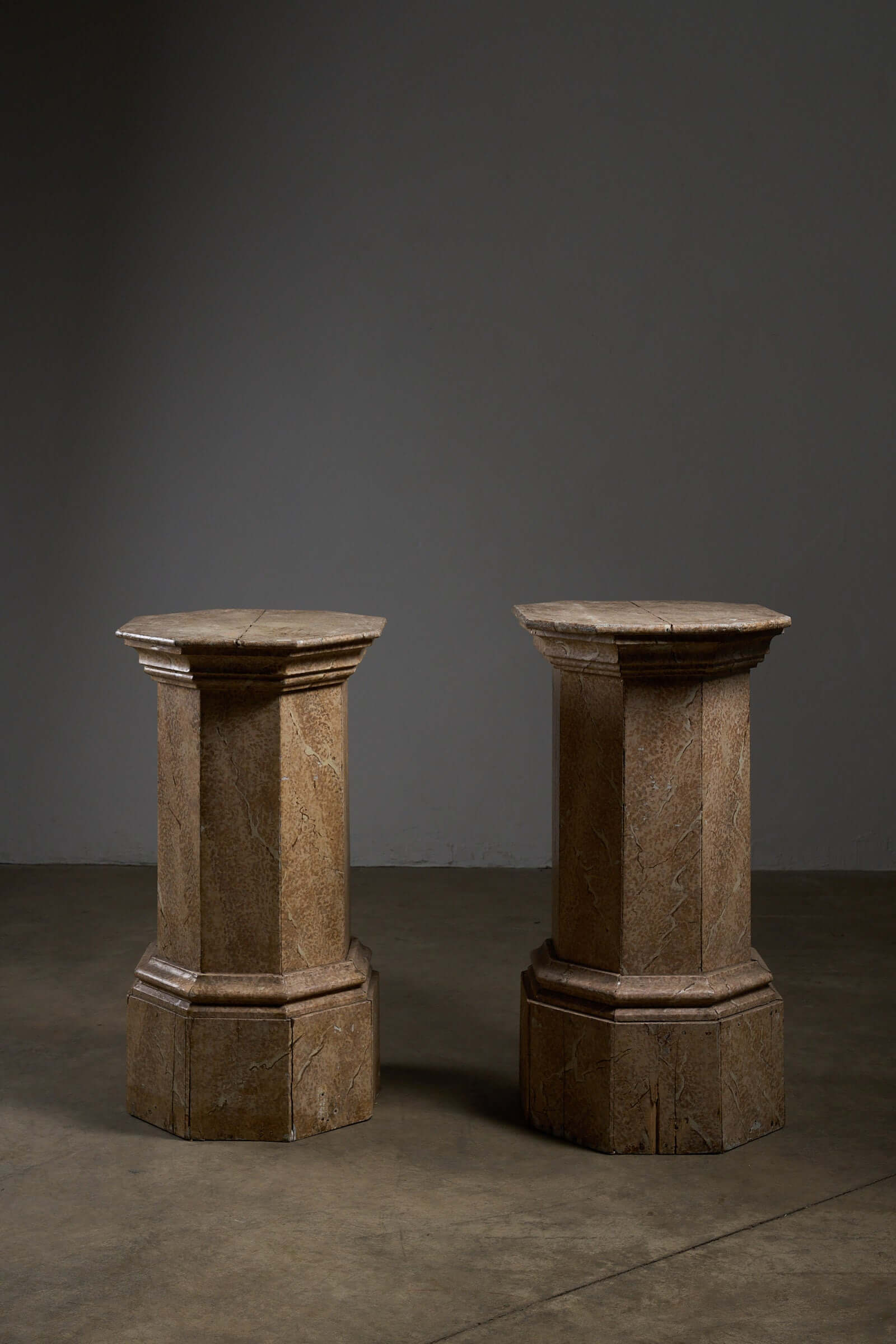 Pair of Antique Faux Marble Octagonal Pedestals showcased in a dimly lit setting, highlighting their unique heights and details.