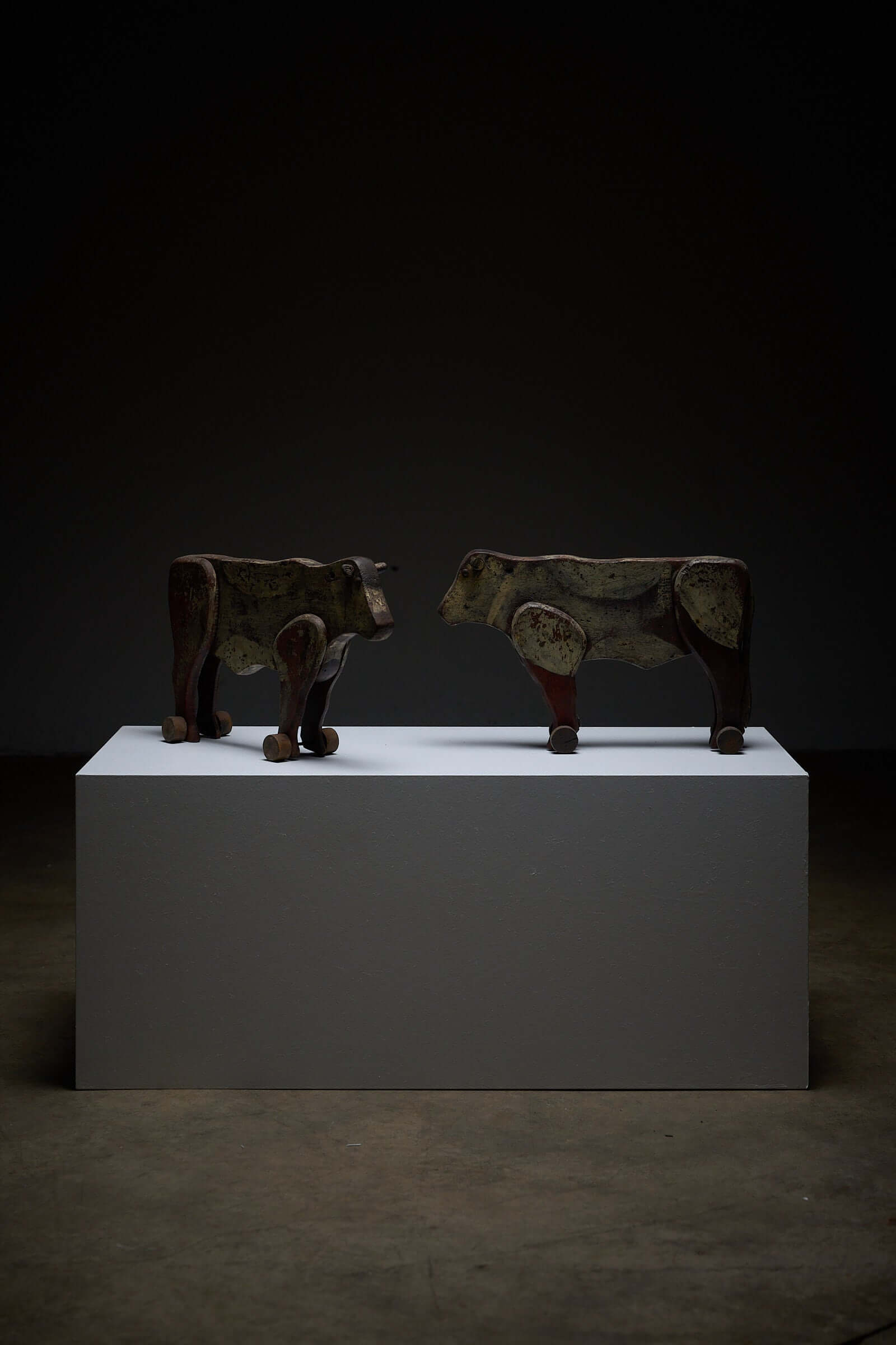 Pair of Antique Articulated Wooden Cow Pull Toys displayed on a pedestal in a dimly lit setting.